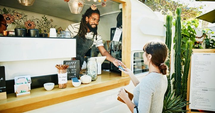 A woman handing her credit card through the window of a food truck to the owner standing inside wearing an apron.