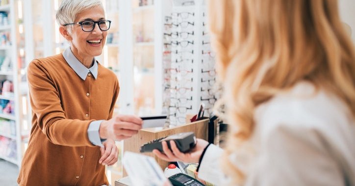 A smiling woman tapping her credit card on a payment terminal being held out by an employee at a glasses store.