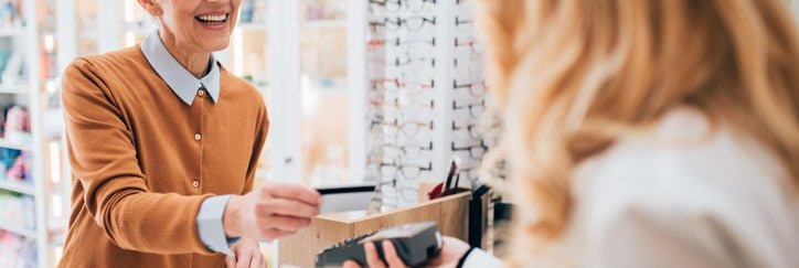 A smiling woman tapping her credit card on a payment terminal being held out by an employee at a glasses store.