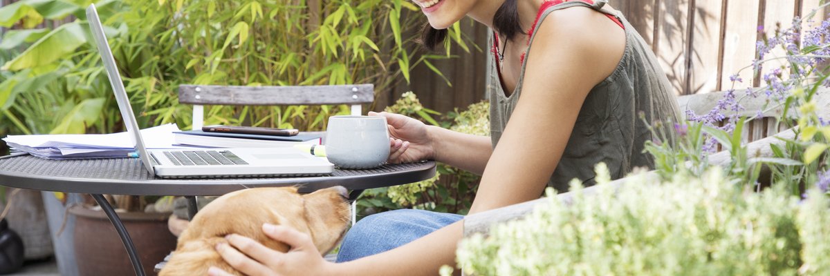 A person petting a dog while sitting outside at a patio table with an open laptop and a cup of coffee.