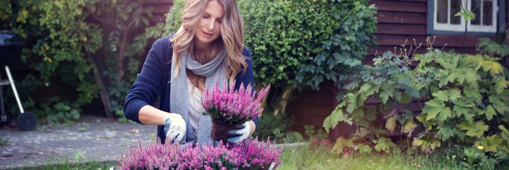 A woman planting flowers in a large pot in her yard with her house in the background.
