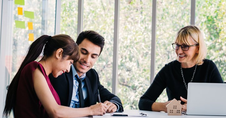 A smiling woman signing documents while sitting at a table with her husband and their realtor.