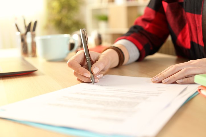 A woman's hands signing paperwork on a desk in a sunny business office.
