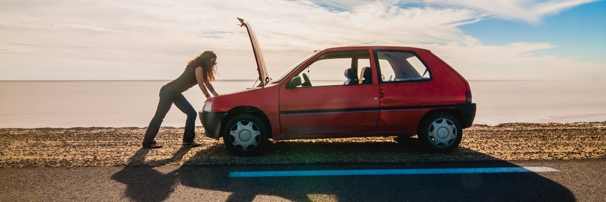woman standing on the side of the road looking under the hood of her car.