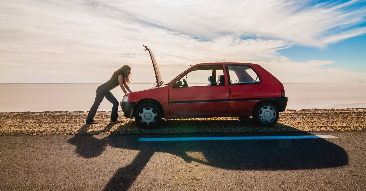 woman standing on the side of the road looking under the hood of her car.