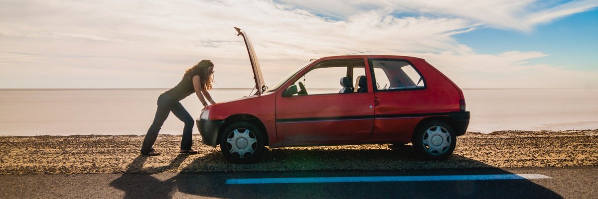 woman standing on the side of the road looking under the hood of her car.