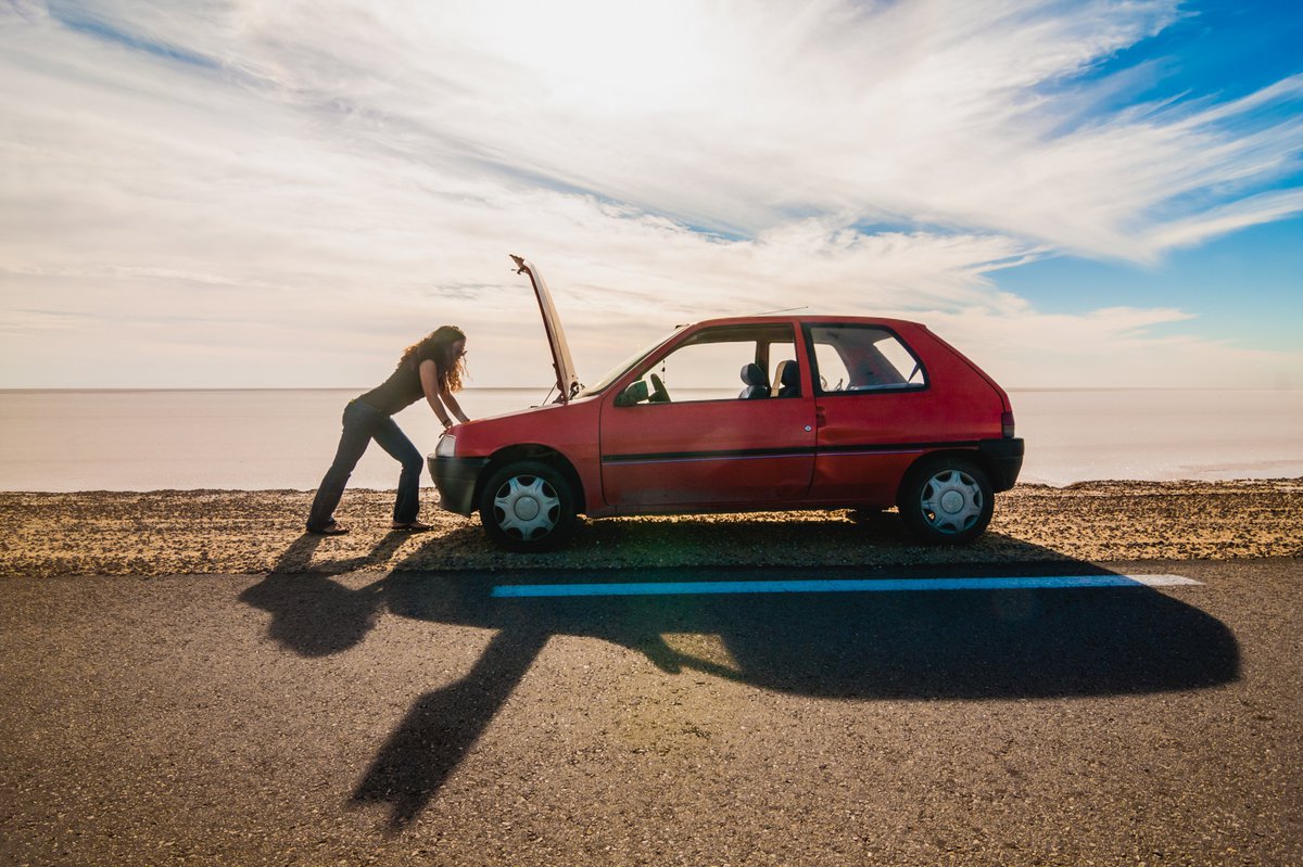 woman standing on the side of the road looking under the hood of her car.