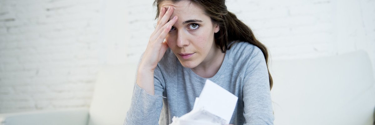 Woman sitting at table with calculator and documents spread out, holding documents in one hand and clutching her head with the other