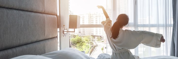 A woman wearing a bathrobe and stretching her arms as she sits on a hotel bed facing the window.