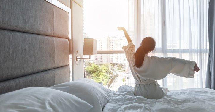 A woman wearing a bathrobe and stretching her arms as she sits on a hotel bed facing the window.
