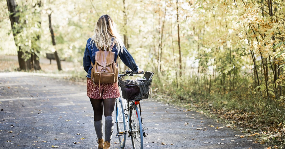 A woman wearing a backpack and walking her bike along a tree-lined path in a park.