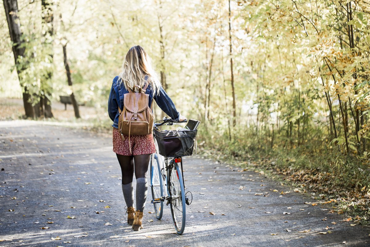A woman wearing a backpack and walking her bike along a tree-lined path in a park.