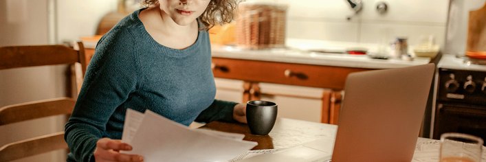 A woman sitting at her kitchen table reviewing paper bills with a laptop open in front of her.