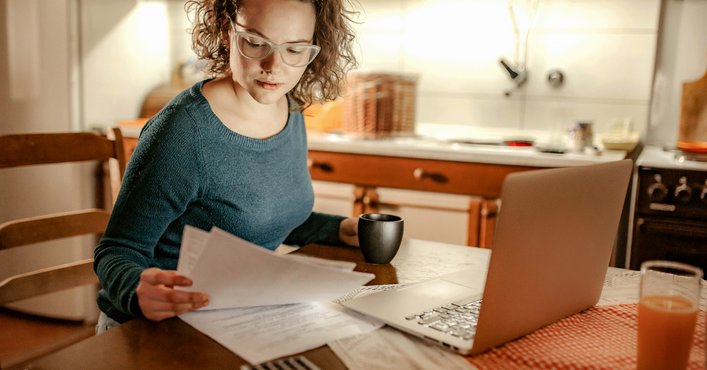A woman sitting at her kitchen table reviewing paper bills with a laptop open in front of her.