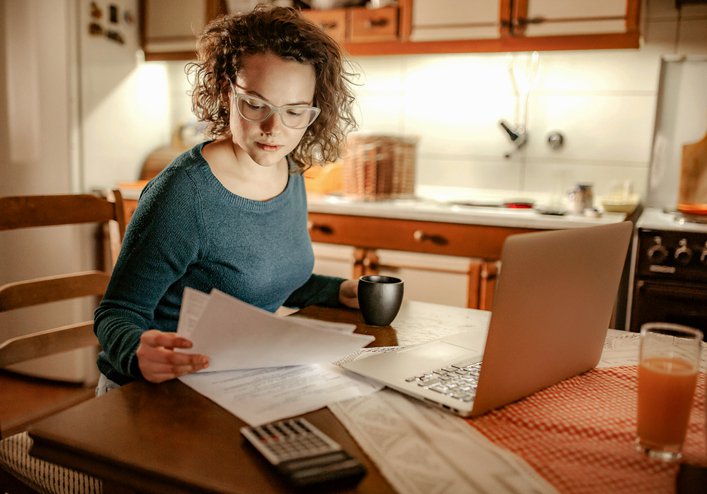 A woman sitting at her kitchen table reviewing paper bills with a laptop open in front of her.
