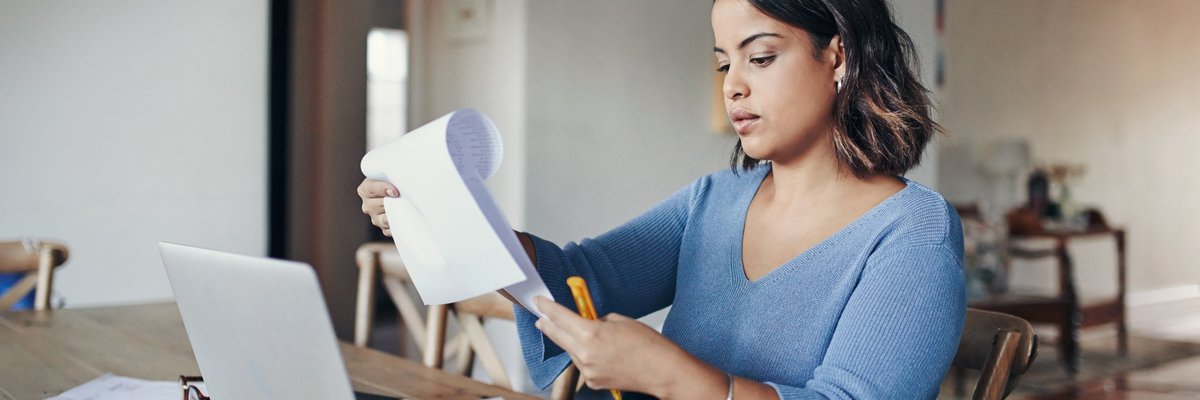 A woman looking over her bills with a laptop and notepad at her kitchen table.