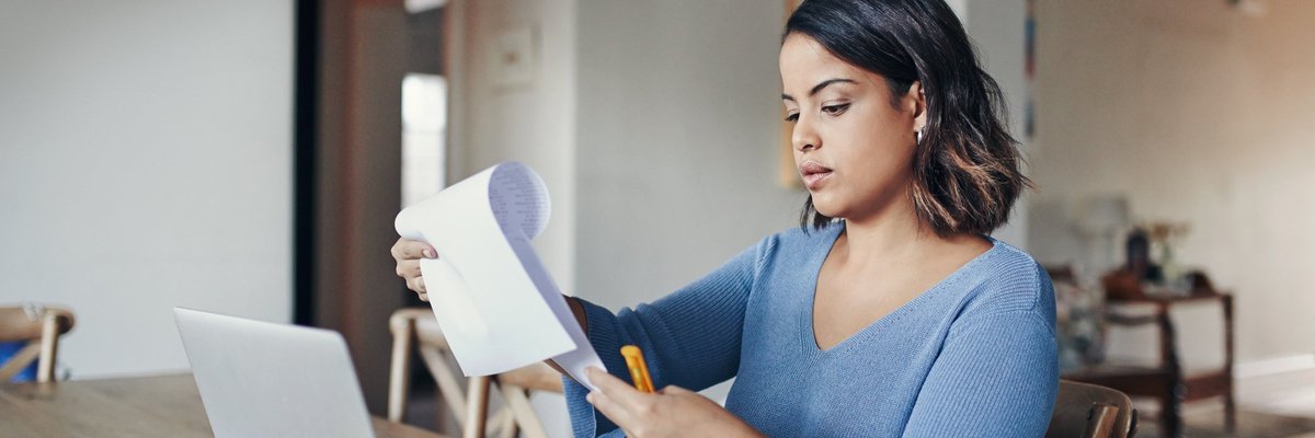 A woman looking over her bills with a laptop and notepad at her kitchen table.