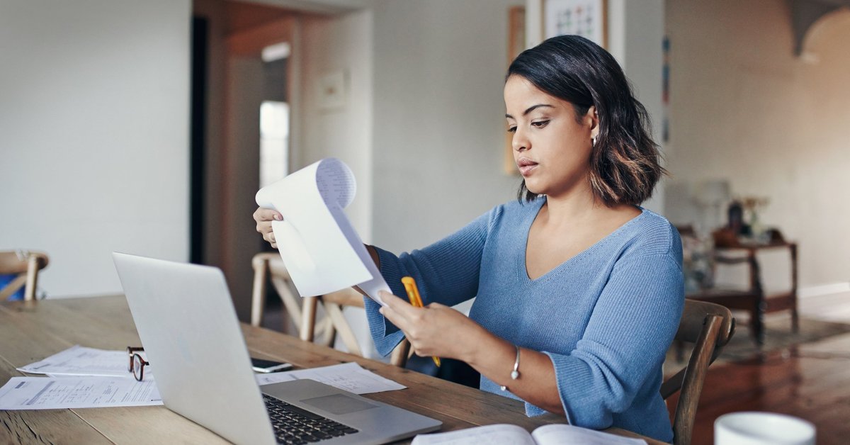 A woman looking over her bills with a laptop and notepad at her kitchen table.