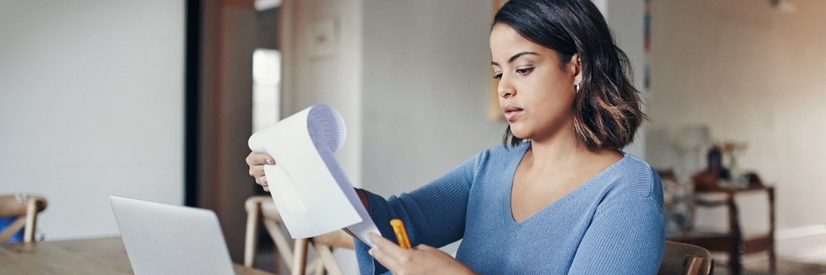 A woman sitting at a dining table with paperwork and a laptop.