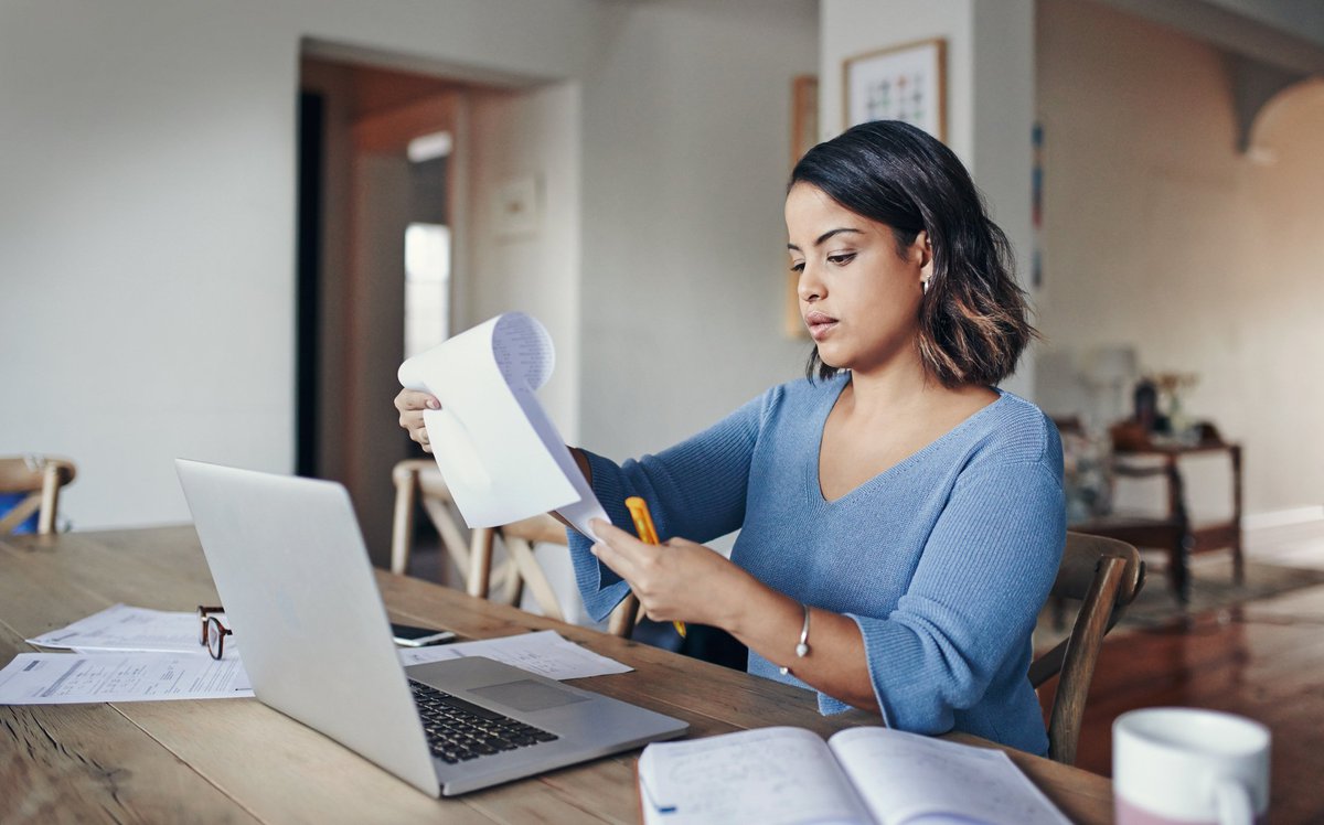 A woman looking over her bills with a laptop and notepad at her kitchen table.