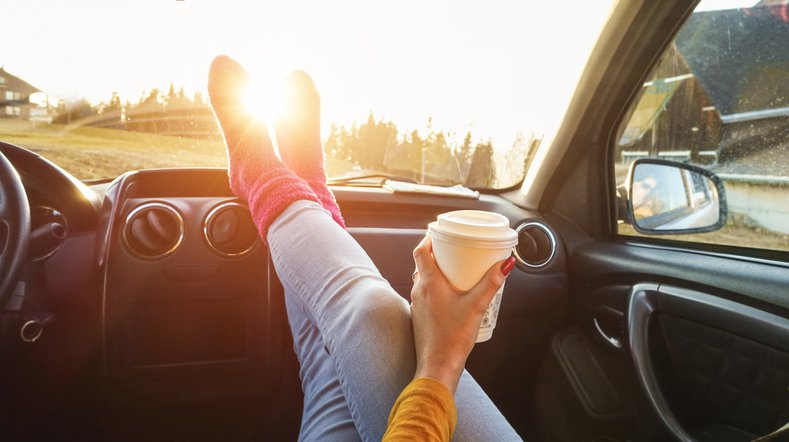 A woman holding a coffee and sitting in the passenger seat of a car pointed toward the sun with her feet resting on the dashboard.