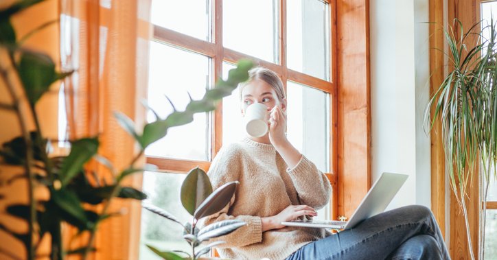 A woman sitting in a sunny window seat at home while drinking coffee and typing on her laptop.