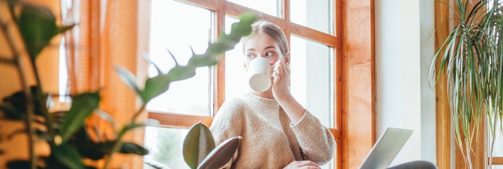 A woman sitting in a sunny window seat at home while drinking coffee and typing on her laptop.