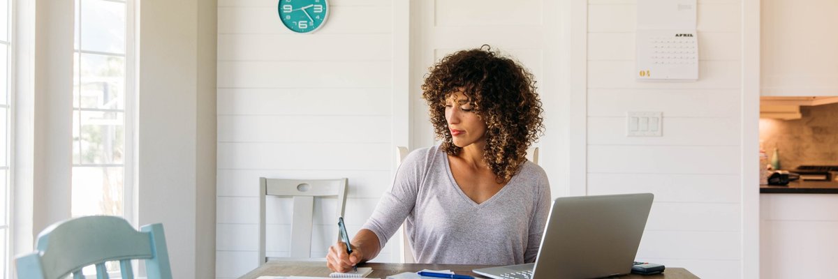 A woman sitting at a sunny kitchen table with her laptop and notes in front of her.