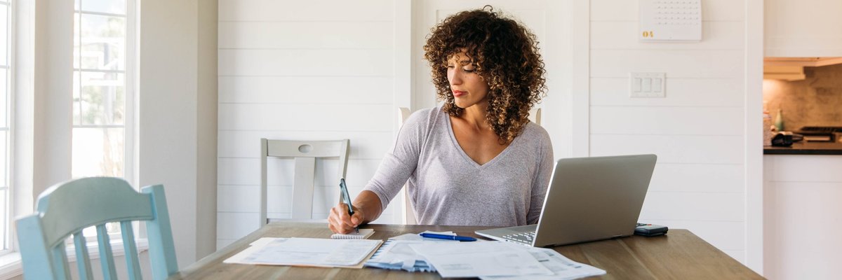 A woman making a budget in a notebook with her laptop open at the kitchen table.