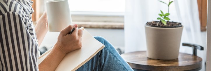 A woman sitting in front of a window at home while writing in a notebook and drinking coffee.