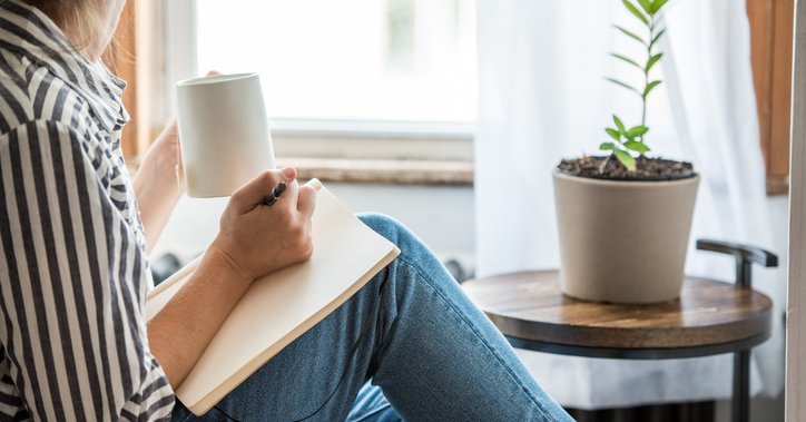 A woman sitting in front of a window at home while writing in a notebook and drinking coffee.