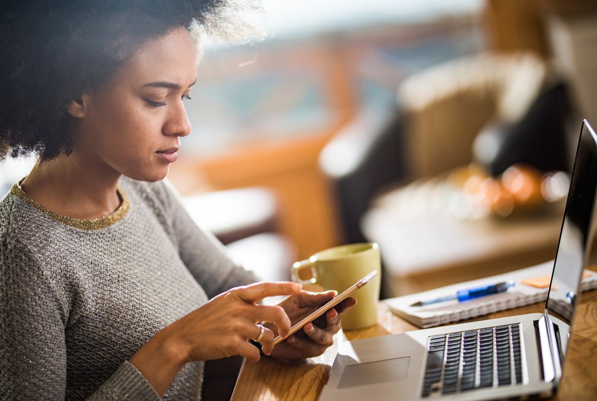 A woman dialing a number on her phone and sitting at a table with her laptop.