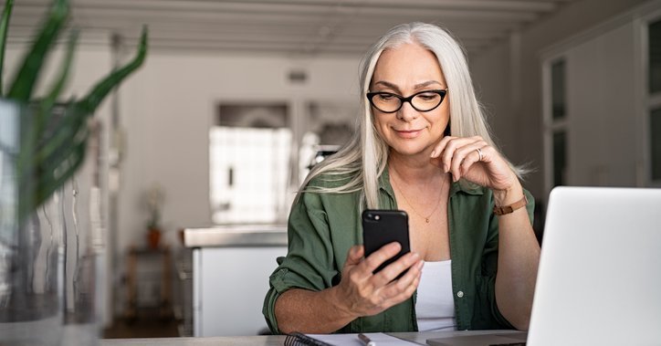 A smiling woman looking at the phone in her hand while sitting at a desk in her living room with her laptop open in front of her.