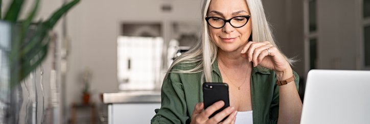 A smiling woman looking at the phone in her hand while sitting at a desk in her living room with her laptop open in front of her.