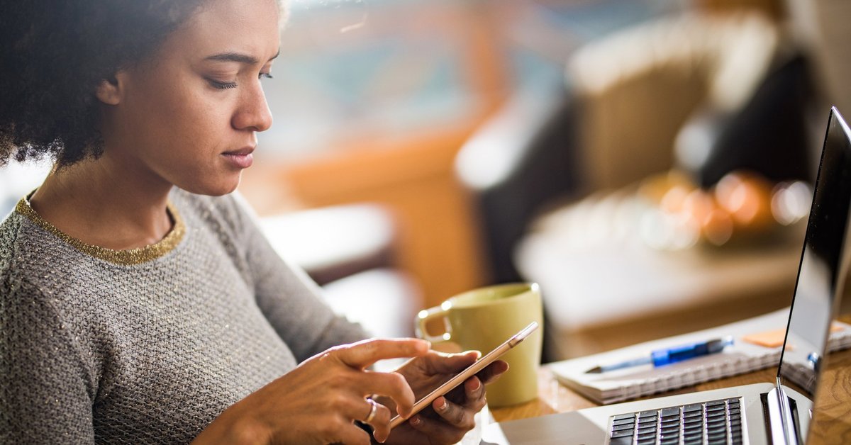 A woman dialing a number on her phone and sitting at a table with her laptop.