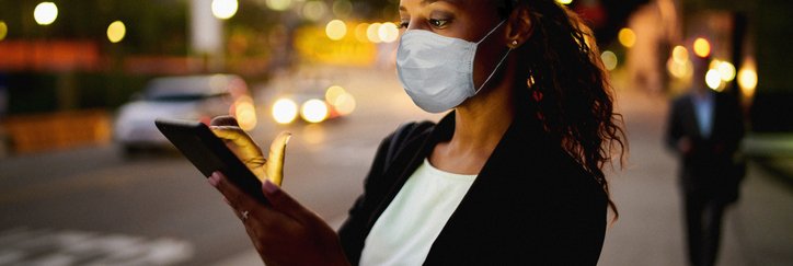 A woman in a mask looks at her phone on a street at dusk.