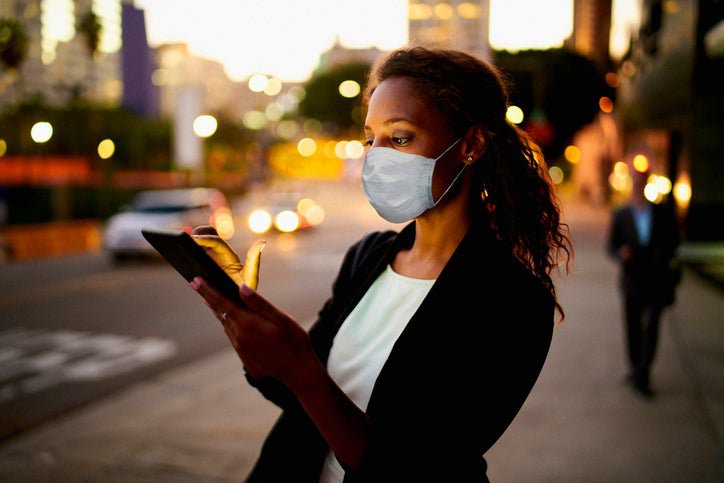 A woman in a mask looks at her phone on a street at dusk.
