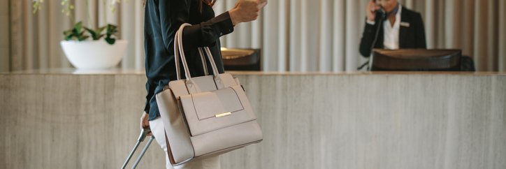 A smiling woman talking on her phone while walking with her suitcase into the lobby of a hotel.