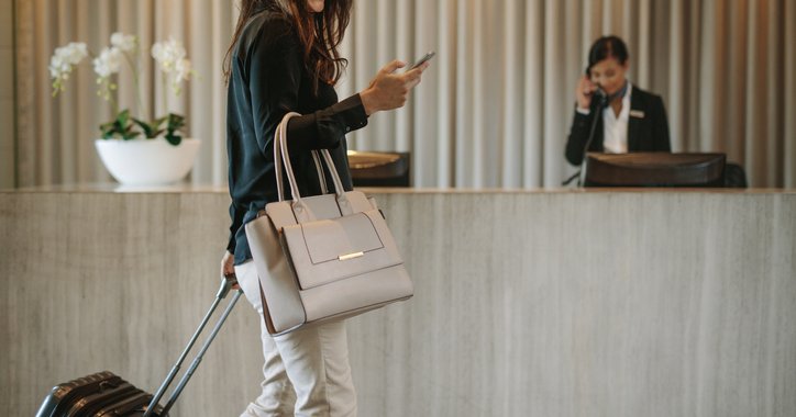 A smiling woman talking on her phone while walking with her suitcase into the lobby of a hotel.