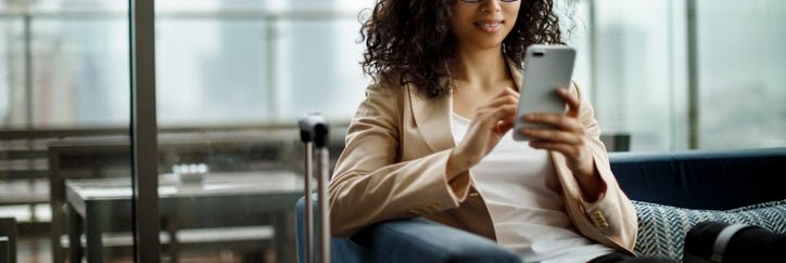 A woman sitting in an airport lounge next to her suitcase and looking at her phone.