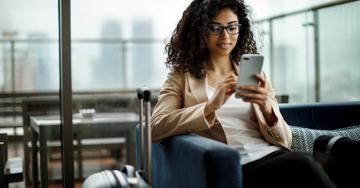 A woman sitting in an airport lounge next to her suitcase and looking at her phone.