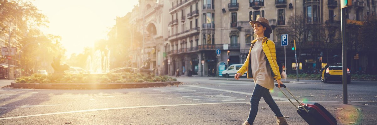 A woman with a suitcase crossing a street in a foreign city with the. sun behind her.