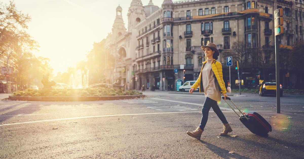 A woman with a suitcase crossing a street in a foreign city with the. sun behind her.