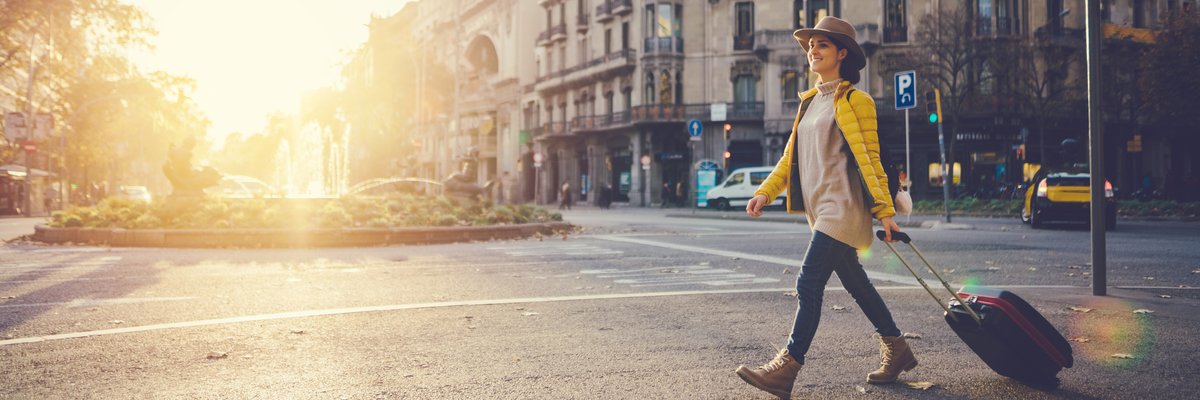 A woman with a suitcase crossing a street in a foreign city with the. sun behind her.