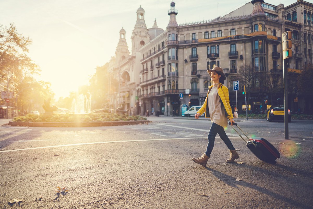 A woman with a suitcase crossing a street in a foreign city with the. sun behind her.