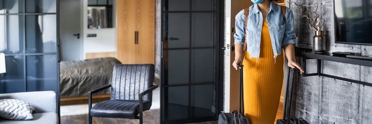 A woman wearing a medical mask and wheeling her two suitcases into a nice hotel room.