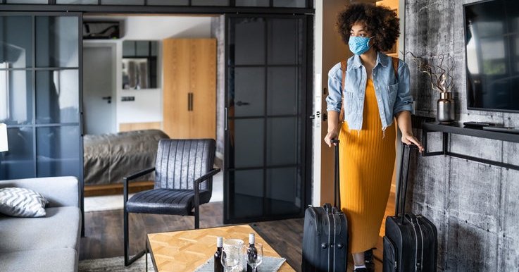 A woman wearing a medical mask and wheeling her two suitcases into a nice hotel room.