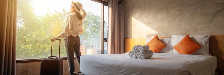 A woman standing next to her suitcase and looking out the window of her sunny hotel room.