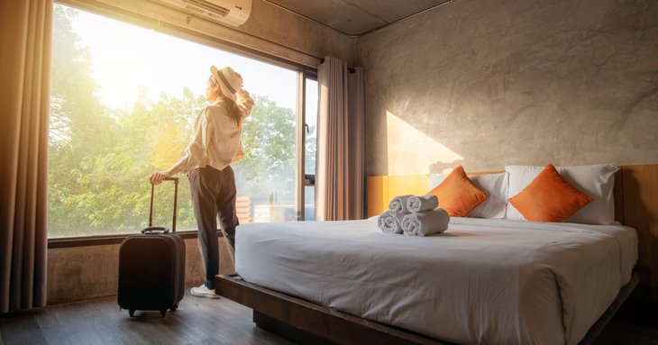 A woman standing next to her suitcase and looking out the window of her sunny hotel room.