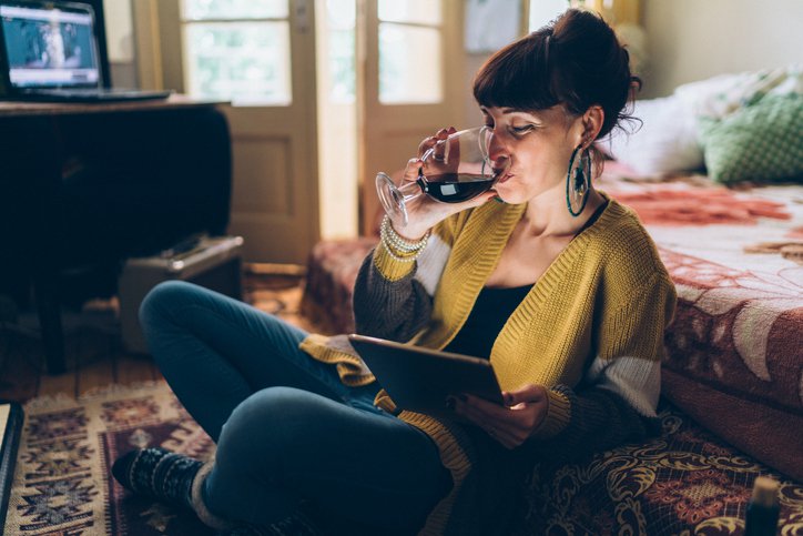 A woman sitting on the floor drinking wine while watching a movie on her tablet.